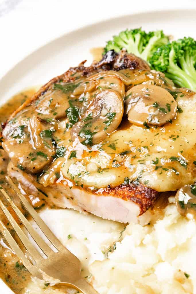 A plate of smothered pork chops topped with mushroom gravy, accompanied by creamy mashed potatoes and steamed broccoli. A fork rests on the side, capturing a homely and appetizing meal.