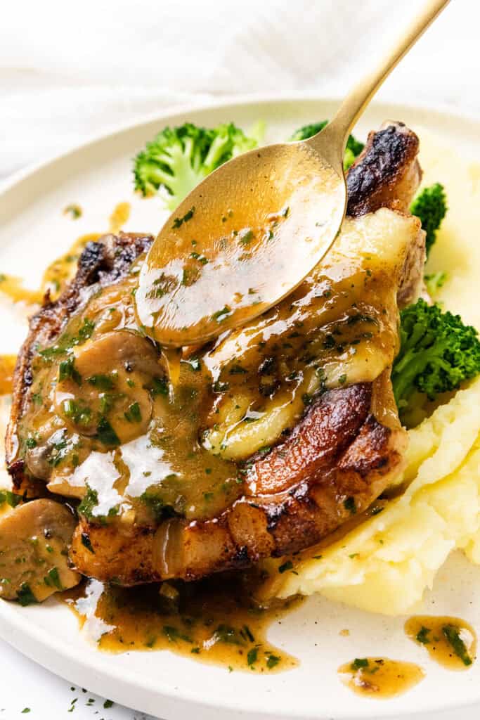 A golden spoon drizzles mushroom sauce over a smothered pork chop, perfectly grilled and garnished with fresh parsley. This delectable dish is served alongside creamy mashed potatoes and vibrant broccoli florets.