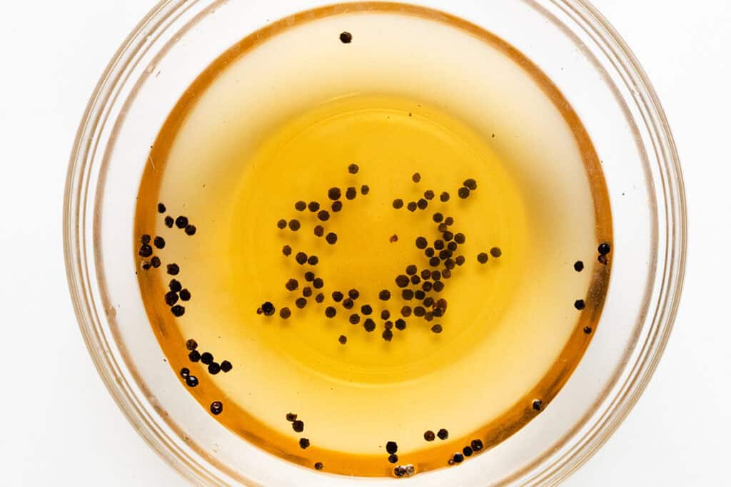 A clear glass bowl filled with amber liquid, possibly oil or syrup, with small black round objects scattered on the surface—like the perfect glaze for smothered pork chops. The bowl rests on a plain white background, inviting culinary inspiration.