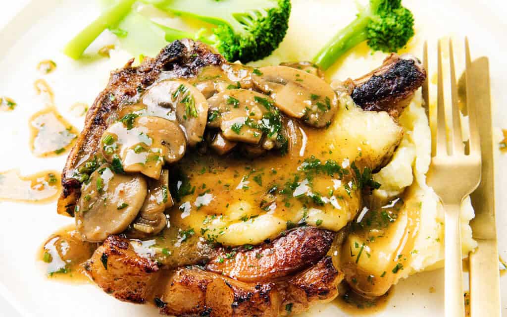 A close-up of a plate features smothered pork chops topped with sliced mushrooms and brown gravy. Garnished with chopped parsley, it sits alongside creamy mashed potatoes and steamed broccoli. A fork and knife rest on the right side for easy dining.