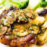 A close-up of a plate features smothered pork chops topped with sliced mushrooms and brown gravy. Garnished with chopped parsley, it sits alongside creamy mashed potatoes and steamed broccoli. A fork and knife rest on the right side for easy dining.