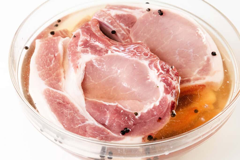 In a glass bowl, two pieces of raw pork are marinating in a flavorful liquid, reminiscent of smothered pork chops. The mixture is speckled with black peppercorns and aromatic spices, enhancing the enticing pink pork with its delicate white marbling.