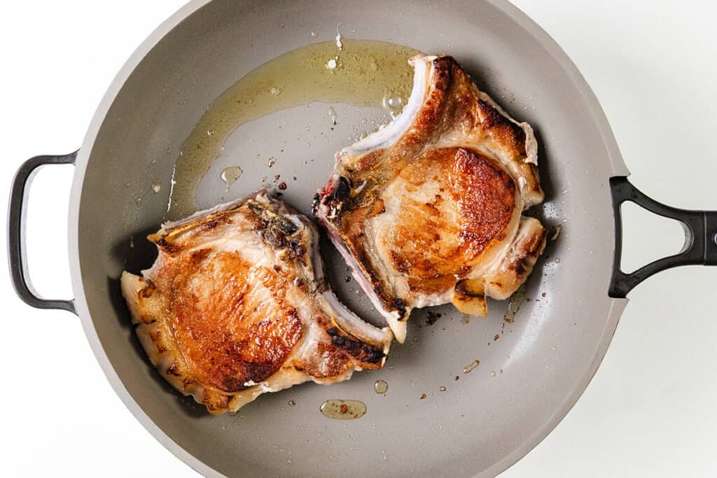 Two browned pork chops sizzle in a gray frying pan with a little oil. Smothered to perfection, the chops have a golden finish with some charred edges, and are positioned on a white background for a minimalist appearance.