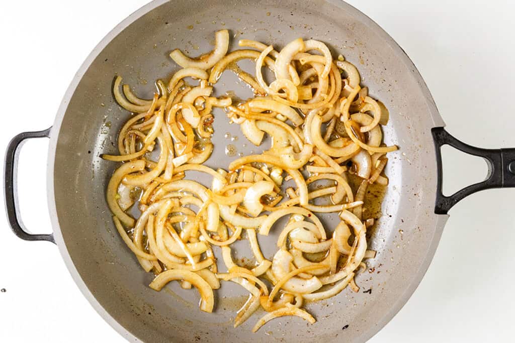 Sliced onions are being sautéed in a grey frying pan, gently browned and spread across the surface. The pan, which features two black handles, is perfect for creating delicious smothered pork chops against a plain white background.