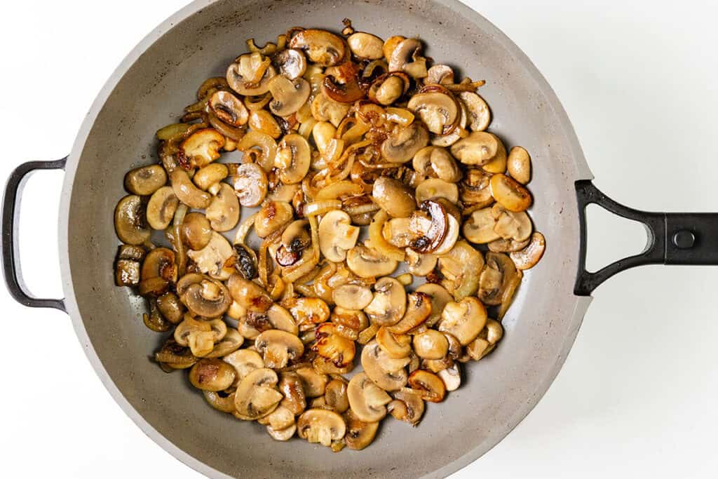 A gray frying pan filled with sautéed sliced mushrooms, lightly browned and glistening with oil, complements the smothered pork chops set against a white background. The mushrooms are evenly distributed across the pan, showcasing their varied shades of brown.