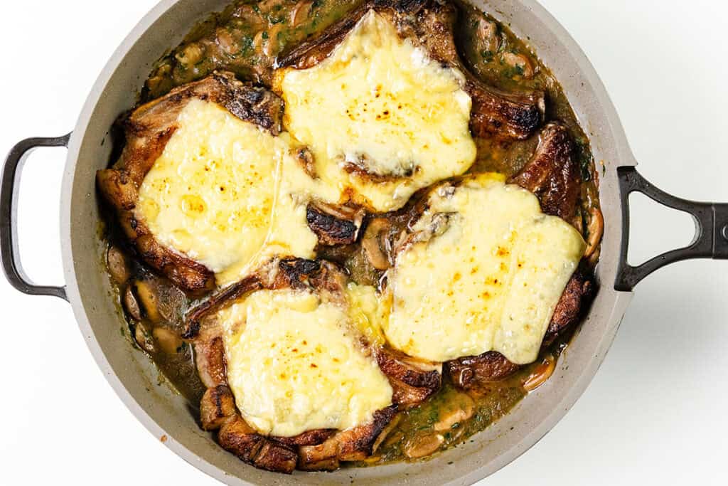 A pan of smothered pork chops topped with melted cheese, nestled in a rich mushroom and onion sauce. The sauce is brown and flavorful, with visible slices of mushrooms and onions enveloping the cooked pork chops.