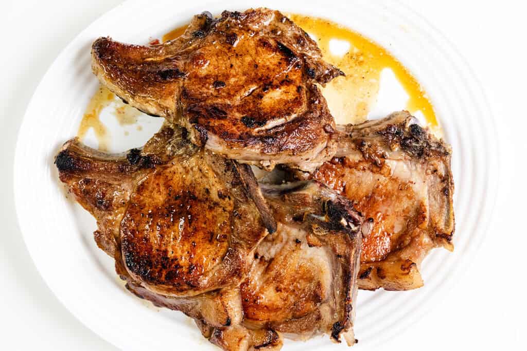 Three grilled pork chops, perfectly browned with visible grill marks and juices pooling around them, evoke the flavors of smothered pork chops. The succulent meat rests invitingly on a pristine white plate.