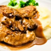 A plate featuring smothered pork chops topped with sautéed mushrooms and gravy, served alongside smooth mashed potatoes and a small portion of steamed broccoli.