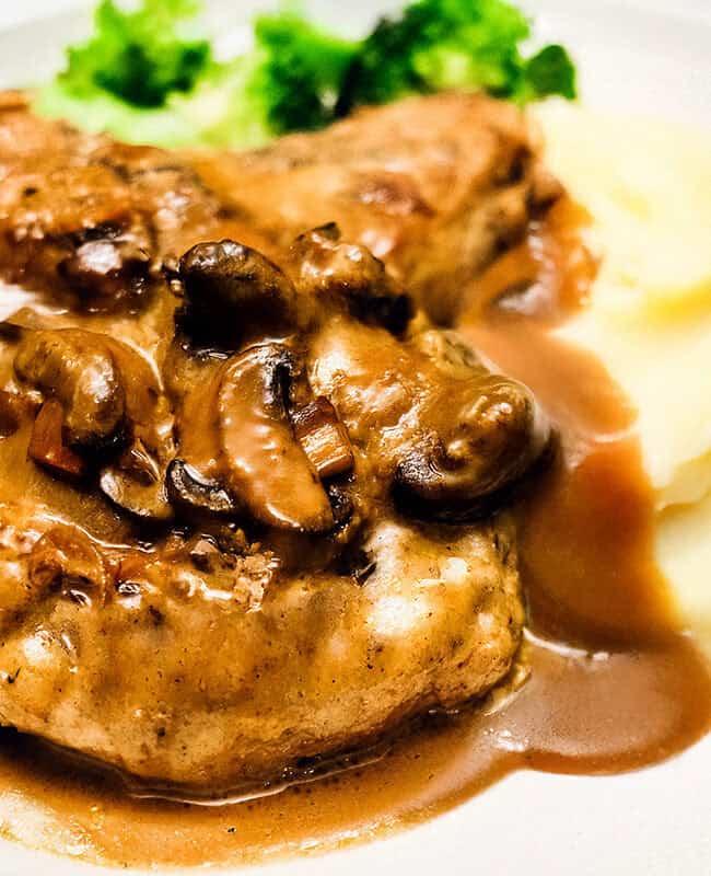 A plate featuring smothered pork chops topped with sautéed mushrooms and gravy, served alongside smooth mashed potatoes and a small portion of steamed broccoli.