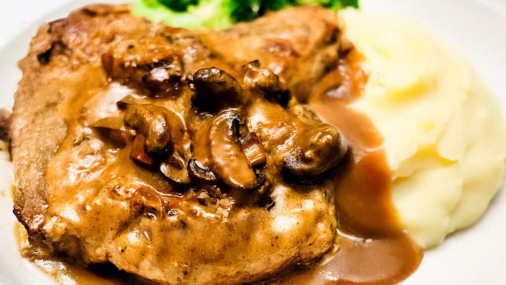 A plate of mashed potatoes and pork chops smothered in mushroom gravy, served with a side of steamed broccoli, reminiscent of classic smothered pork chops.