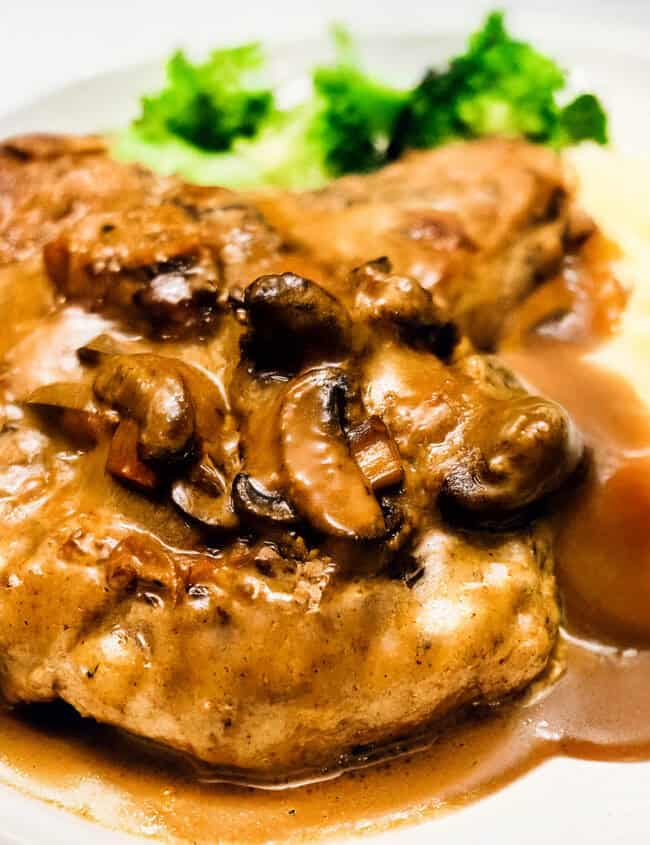 A dish featuring a chicken breast reminiscent of smothered pork chops, bathed in mushroom gravy, served with mashed potatoes and a side of steamed broccoli on a white plate. The meal appears freshly cooked and richly flavored.