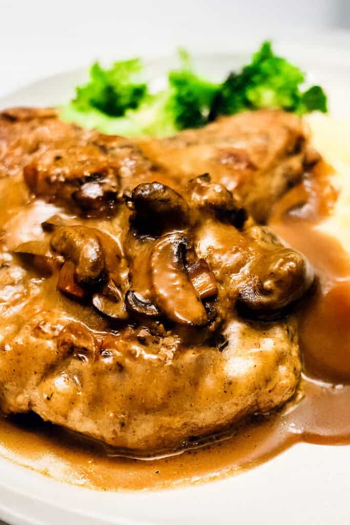 A dish featuring a pork chop bathed in mushroom gravy, served with mashed potatoes and a side of steamed broccoli on a white plate. The meal appears freshly cooked and richly flavored.