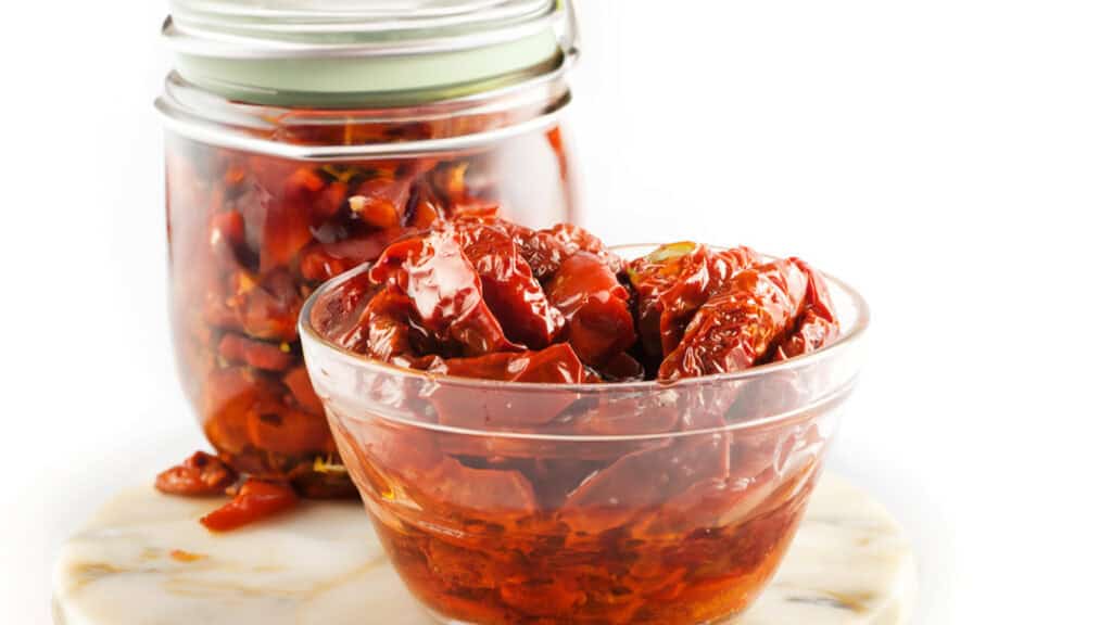 A close up image of sun-dried tomatoes in a bowl with the jar behind it.