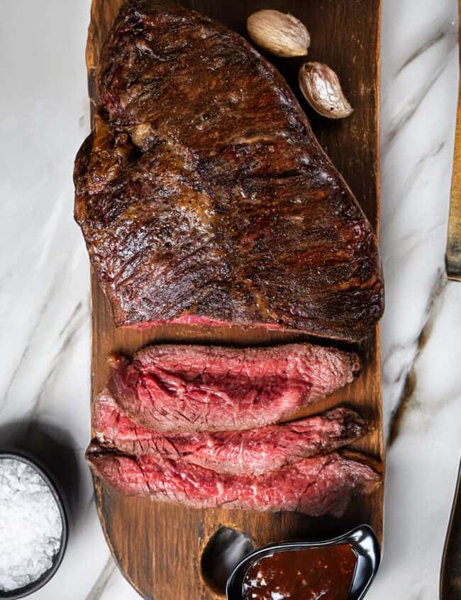 A wooden board showcases a pan fried steak, partially sliced to reveal a rare center, accompanied by whole garlic cloves. Beside the board is a small bowl of coarse salt and a black dish filled with barbecue sauce, all elegantly arranged on a marble surface.