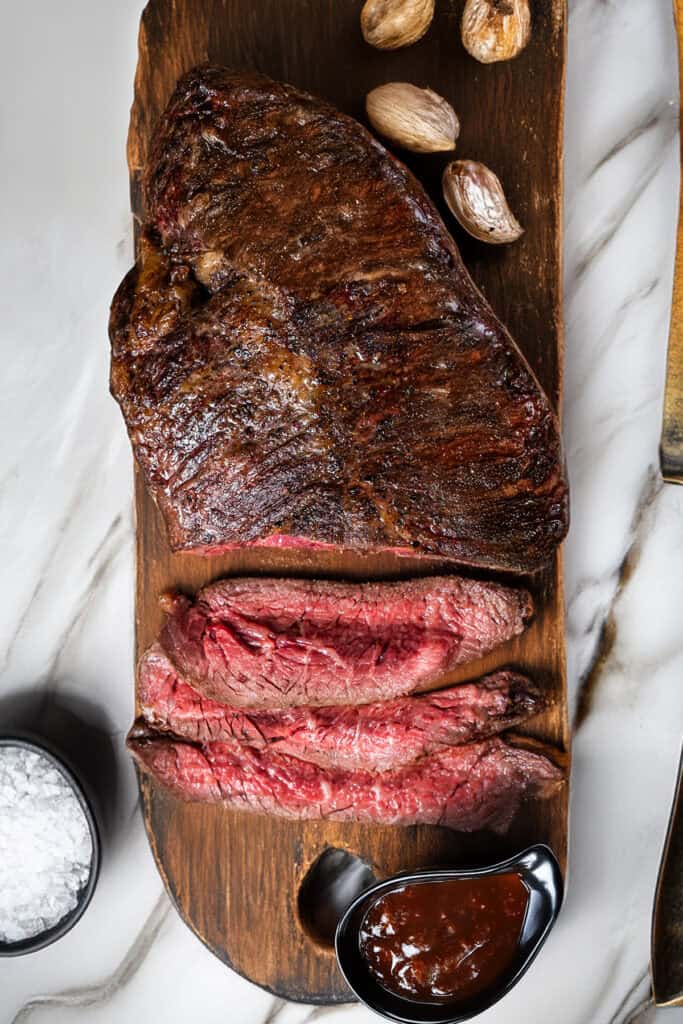 A wooden board showcases a pan fried steak, partially sliced to reveal a rare center, accompanied by whole garlic cloves. Beside the board is a small bowl of coarse salt and a black dish filled with barbecue sauce, all elegantly arranged on a marble surface.