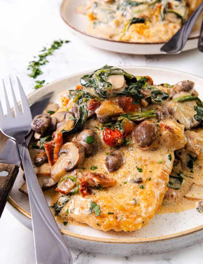 A plate of creamy Tuscan chicken with sun-dried tomatoes, spinach, and mushrooms, served with a fork and knife.