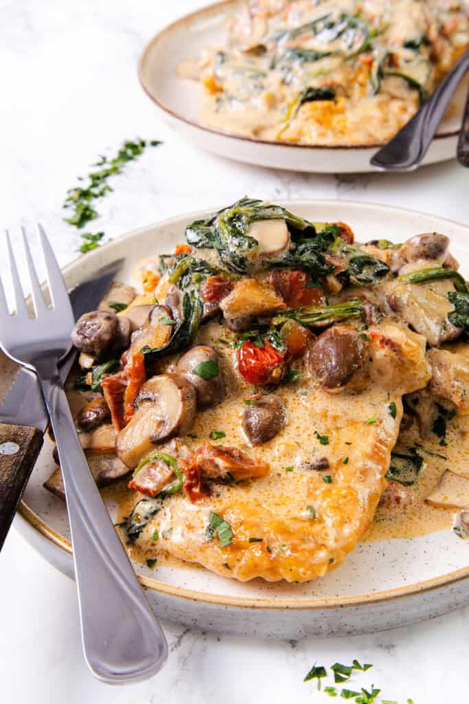 Plate of creamy Tuscan chicken with sun-dried tomatoes, spinach, and mushrooms, served with a fork and knife.