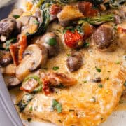 A close up image of a dish of creamy Tuscan chicken with sun-dried tomatoes, spinach, and mushrooms, served with a fork and knife.