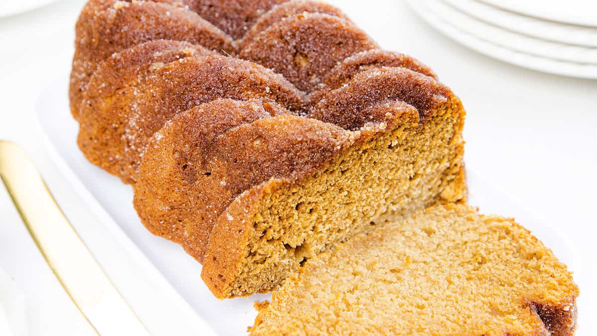 The image shows a loaf of applesauce bread, golden-brown with a sugary crust, partially sliced on a white plate next to a gold-colored knife.