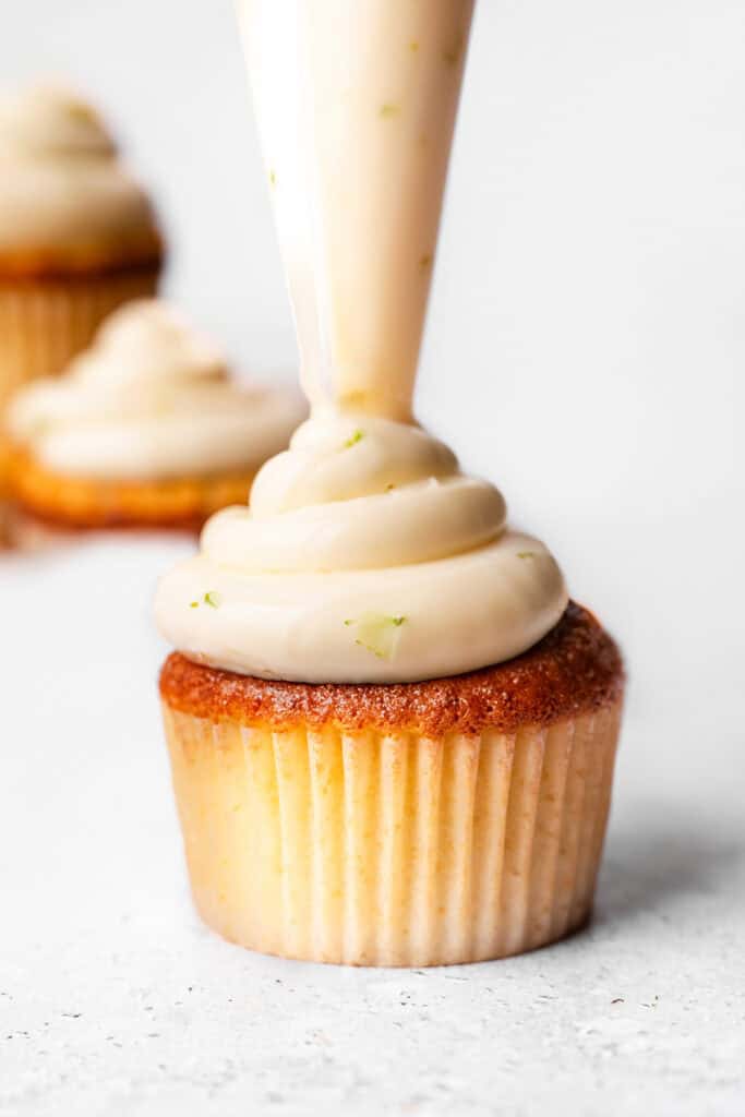 A close-up of a Key Lime Cupcake being frosted with creamy icing, showcasing the smooth and rich texture of the frosting with other frosted cupcakes blurred in the background.