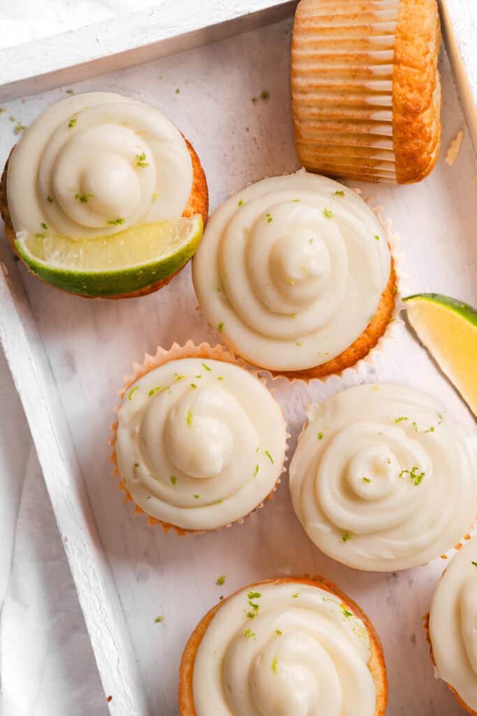 A close-up of Key Lime Cupcakes topped with creamy frosting and garnished with fresh lime zest, displayed in a white tray.