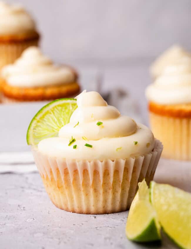 Key lime cupcake with creamy frosting, topped with a lime slice and zest, in a white liner on a marble background.