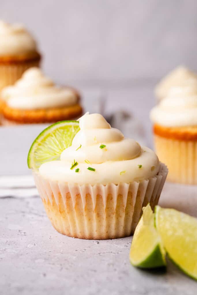 Key lime cupcake with creamy frosting, topped with a lime slice and zest, in a white liner on a marble background.