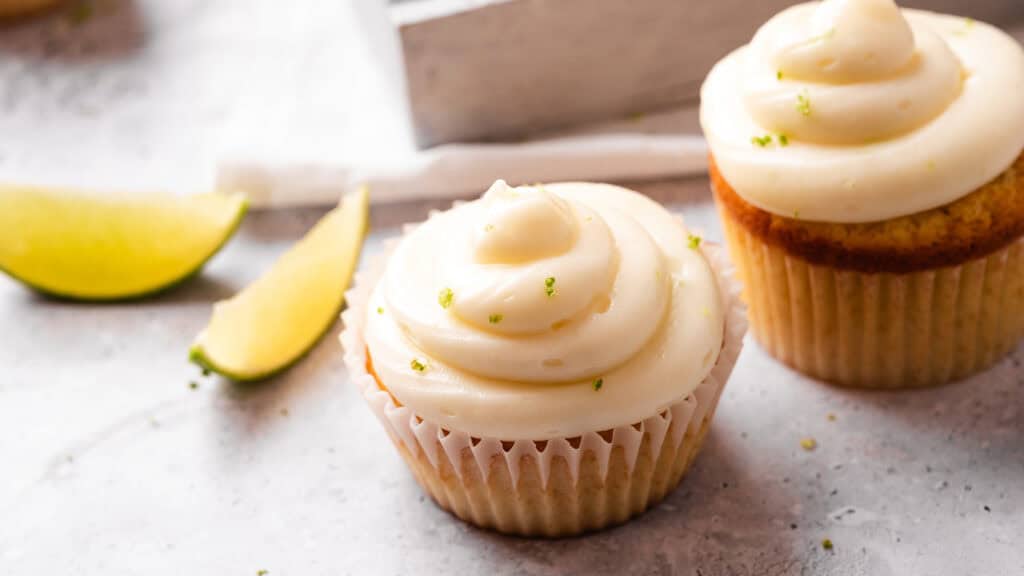 Two Key Lime Cupcakes topped with creamy frosting and garnished with lime zest, with lime wedges in the background.