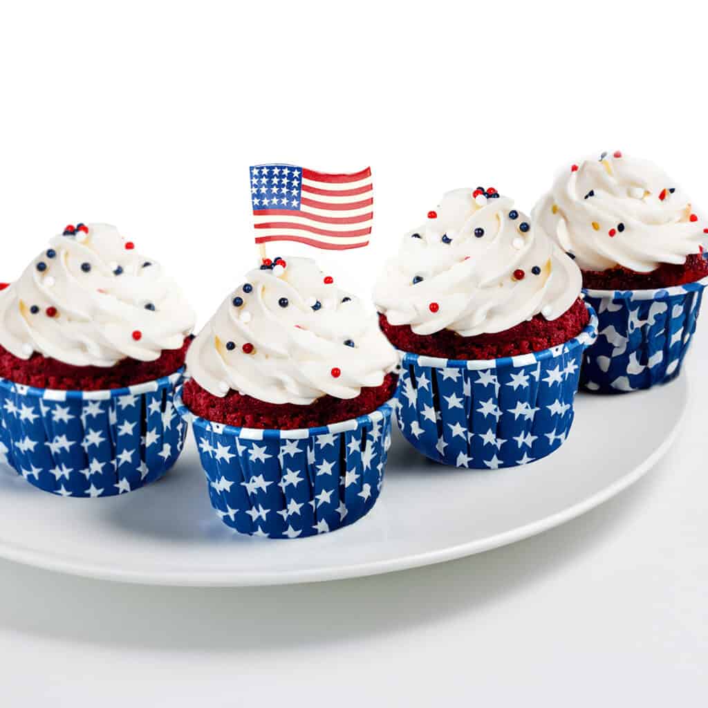 a plate of red velvet 4th of July cupcakes with whipped cream topping and red white and blue sprinkles. One has a mini American flag on top