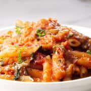 A close-up of a plate of penne pasta topped with a rich red tomato sauce, reminiscent of Pasta Alla Norma. The pasta is garnished with fresh chopped basil and grated cheese, creating a visually appetizing dish. The background is a white marble surface, providing a clean and elegant setting.