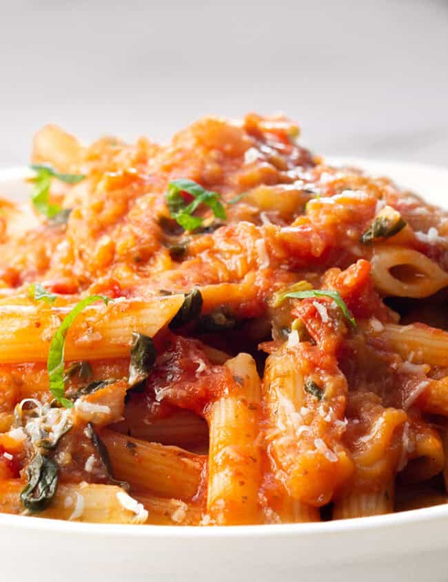 A close-up of a plate of penne pasta topped with a rich red tomato sauce, reminiscent of Pasta Alla Norma. The pasta is garnished with fresh chopped basil and grated cheese, creating a visually appetizing dish. The background is a white marble surface, providing a clean and elegant setting.