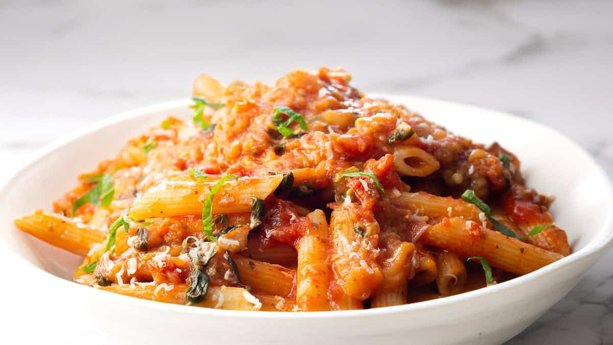 A close-up of a plate of penne pasta topped with a rich red tomato sauce, reminiscent of Pasta Alla Norma. The pasta is garnished with fresh chopped basil and grated cheese, creating a visually appetizing dish. The background is a white marble surface, providing a clean and elegant setting.