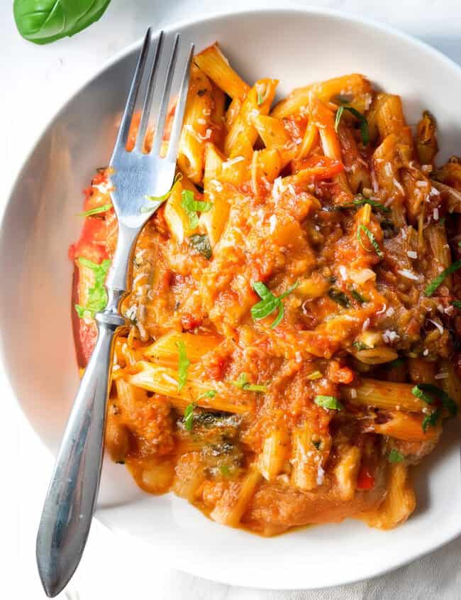 A white bowl filled with penne pasta topped with a chunky tomato sauce and garnished with chopped herbs, reminiscent of Pasta Alla Norma. A fork rests on the side, and fresh basil leaves are seen in the top left corner. The bowl is placed on a white, marble-like surface.