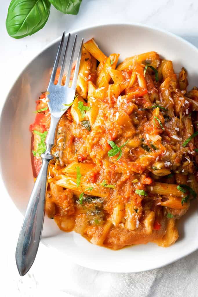 A white bowl filled with penne pasta topped with a chunky tomato sauce and garnished with chopped herbs, reminiscent of Pasta Alla Norma. A fork rests on the side, and fresh basil leaves are seen in the top left corner. The bowl is placed on a white, marble-like surface.