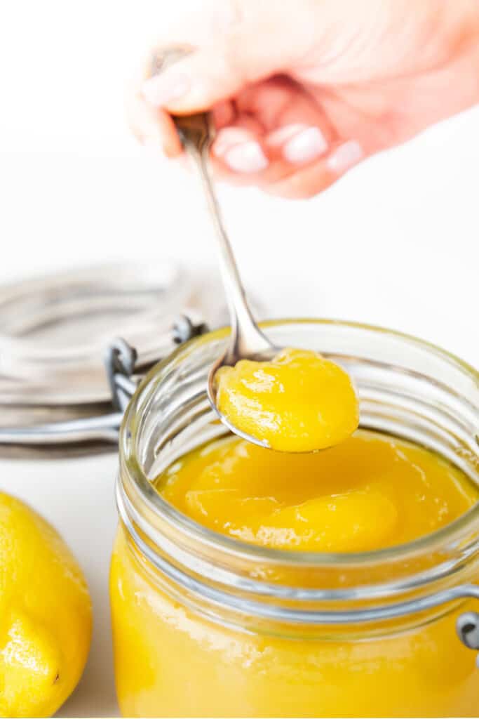 A hand holding a spoonful of lemon curd over a jar of lemon curd.