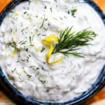 a close up image of a bowl of Tzatziki topped with chopped dill and lemon zest and surrounded by pita bread.