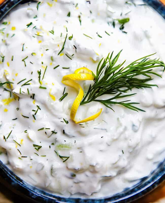 a close up image of a bowl of Tzatziki topped with chopped dill and lemon zest and surrounded by pita bread.