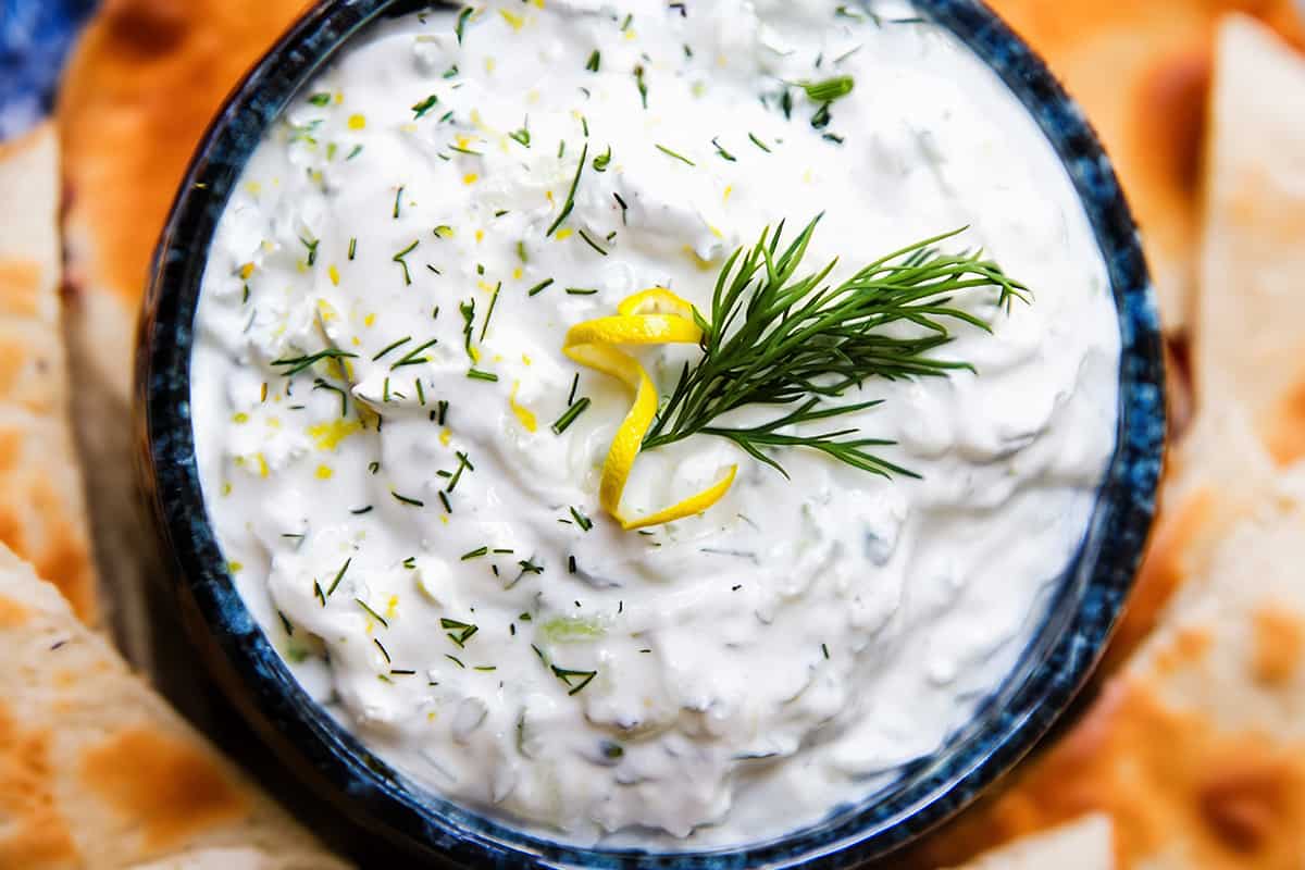 a close up image of a bowl of Tzatziki topped with chopped dill and lemon zest and surrounded by pita bread.