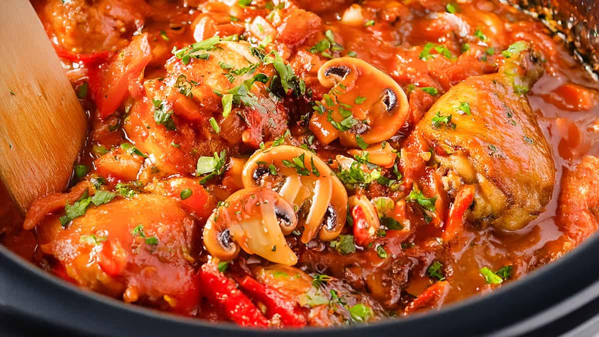 A close-up of a vibrant Chicken Cacciatore dish in a slow cooker featuring chicken pieces stewing in a rich, red tomato sauce with sliced mushrooms and bell peppers. Fresh green herbs are sprinkled on top, and a wooden spoon is partially visible on the side.