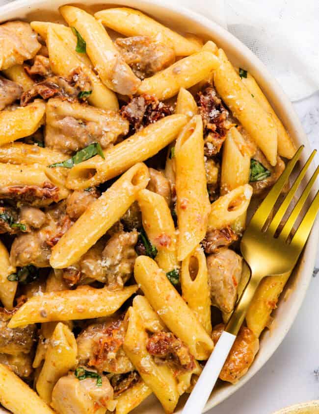 A bowl of sun-dried tomato pasta with creamy penne, tender chicken, and fresh basil. The dish is topped with grated cheese, accompanied by a white napkin and a gold-and-white fork.