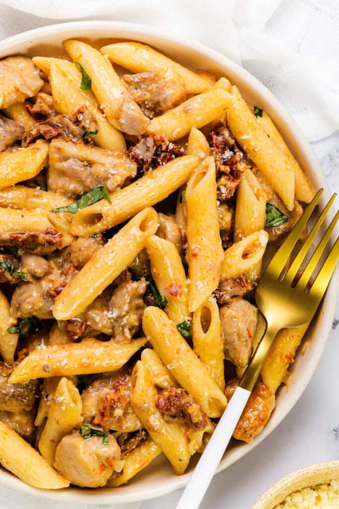 A bowl of sun-dried tomato pasta with creamy penne, tender chicken, and fresh basil. The dish is topped with grated cheese, accompanied by a white napkin and a gold-and-white fork.