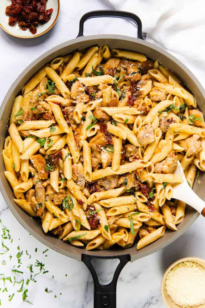 A pan brimming with creamy Sun-Dried Tomato Pasta features succulent chicken pieces and a garnish of fresh basil. A white spoon rests within, surrounded by small bowls of grated cheese and extra sun-dried tomatoes for your culinary delight.