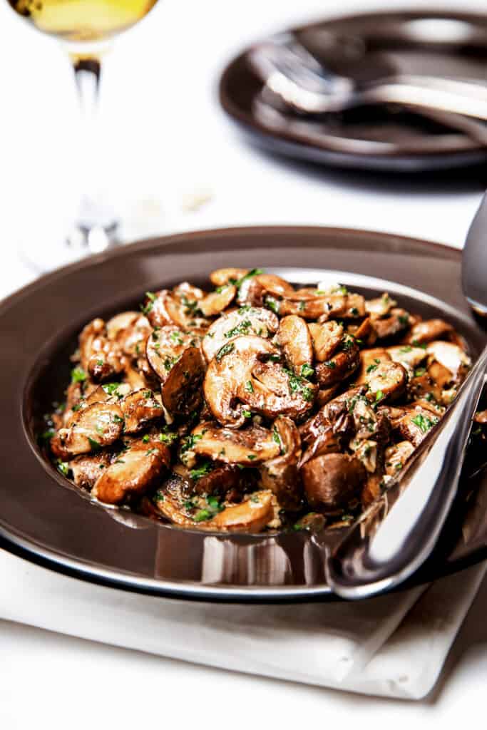 Golden sautéed Mushrooms in a bowl with a serving spoon and a glass of white wine in the background.