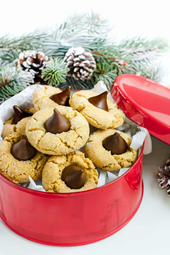 A red cookie tin filled with peanut butter blossom cookies, each topped with a chocolate kiss, set against a backdrop of pine branches and pinecones.