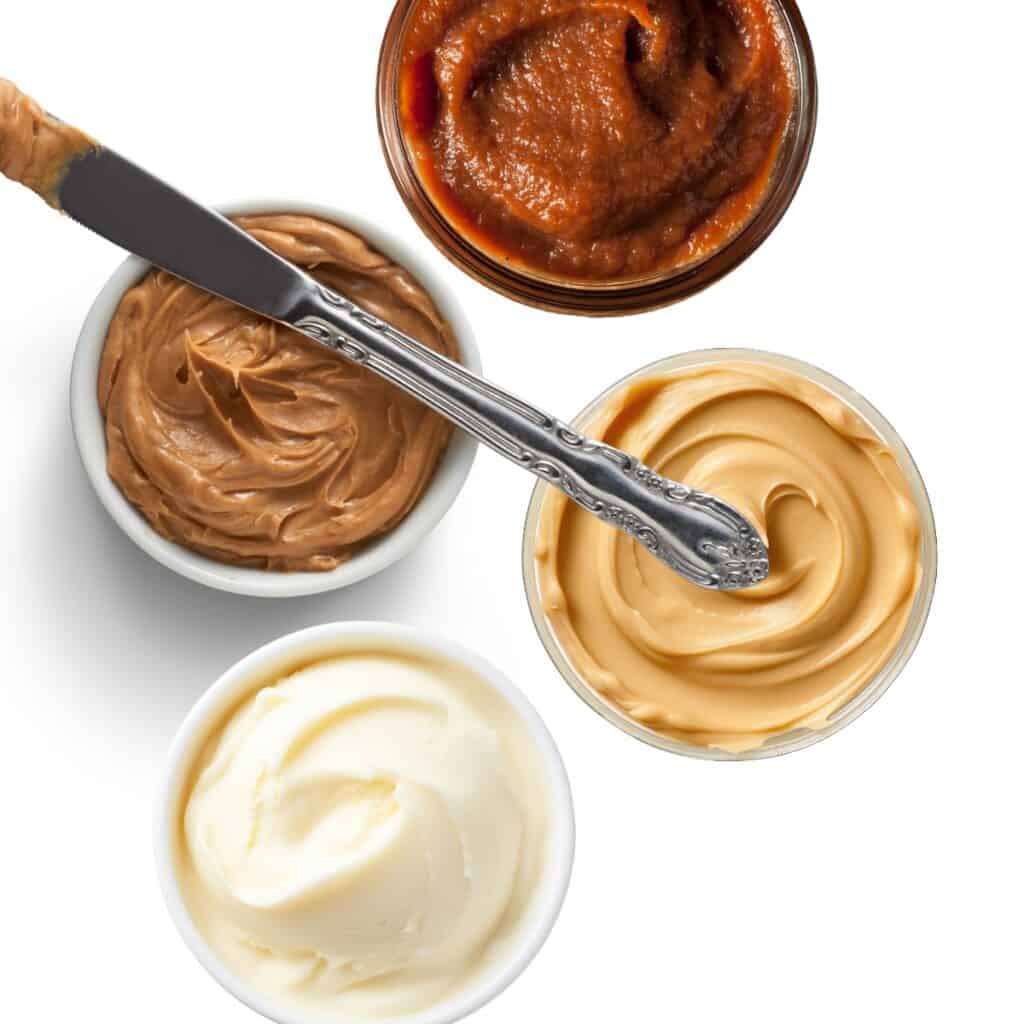 Four small bowls with peanut butter substitutes: one with apple butter, one with sunflower seed butter, cookie butter, and one with soy butter, all on a white background with a knife dipping into the cookie butter.