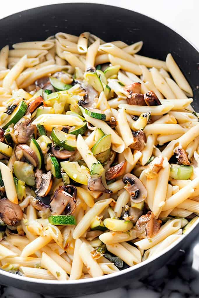 A close-up image of a pan full of Roasted Vegetable Pasta with penne, sliced zucchini, mushrooms, and red onions, showcasing the dish's vibrant and fresh ingredients.