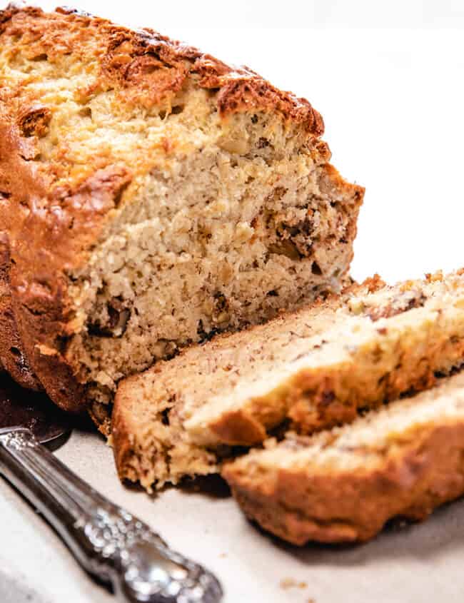 The image features a loaf of banana bread with several slices cut, alongside a serving knife on a stone board.