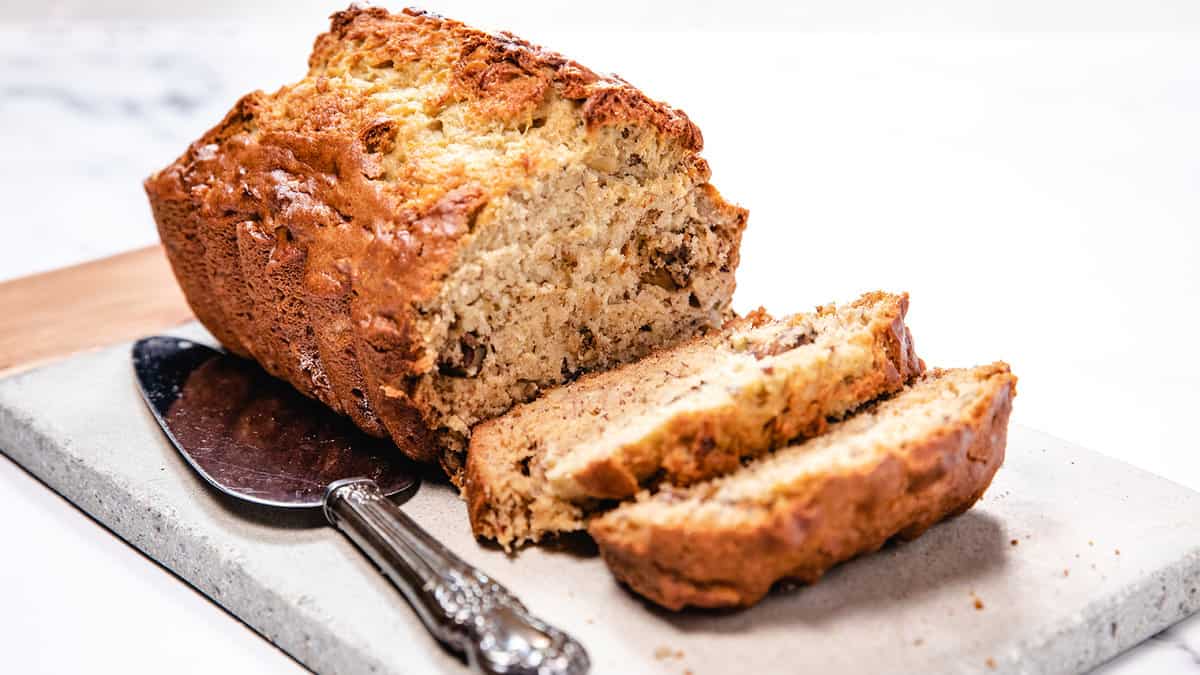 The image features a loaf of banana bread with several slices cut, alongside a serving knife on a stone board.