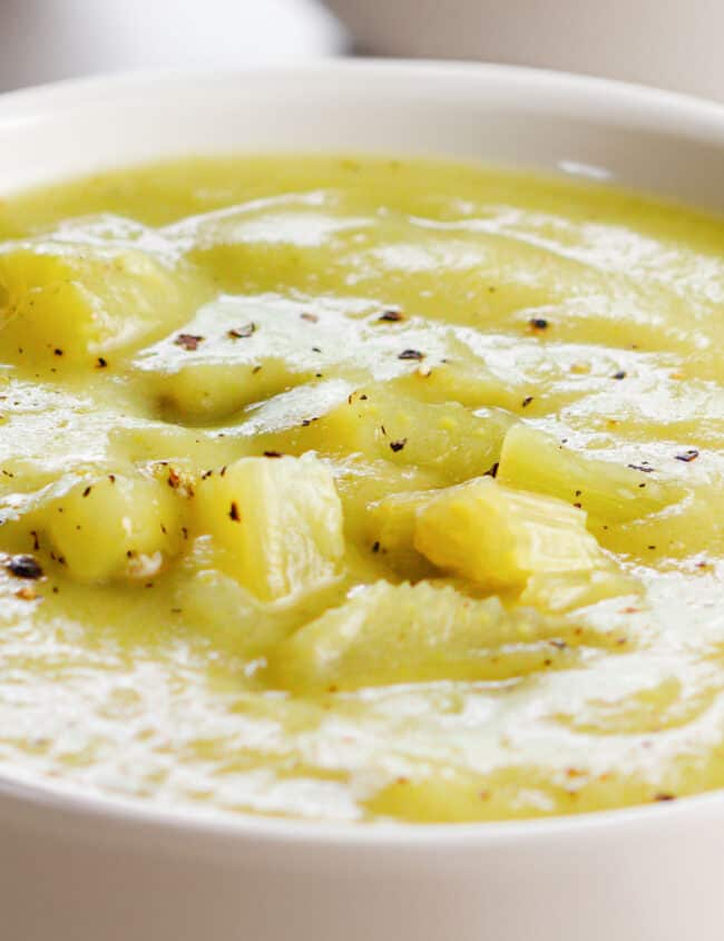 A close up image of a white bowl of creamy celery soup, seasoned with cracked black pepper, with chunks of celery on top.