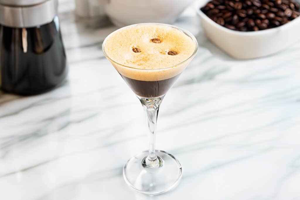 Espresso martini in a clear glass with a creamy foam top garnished with coffee beans on a marble countertop with coffee-making equipment in the background.